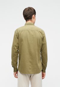 Mann mit kurzen braunen Haaren, der ein olivgrünes, langärmeliges Hemd mit Knöpfen und helle beige Hosen trägt, steht mit dem Rücken zum Betrachter vor einem weißen Hintergrund.