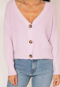 Cardigan tricoté rose clair avec un décolleté en V profond, quatre boutons marron, texture côtelée et hem court, associé à un jean bleu taille haute.