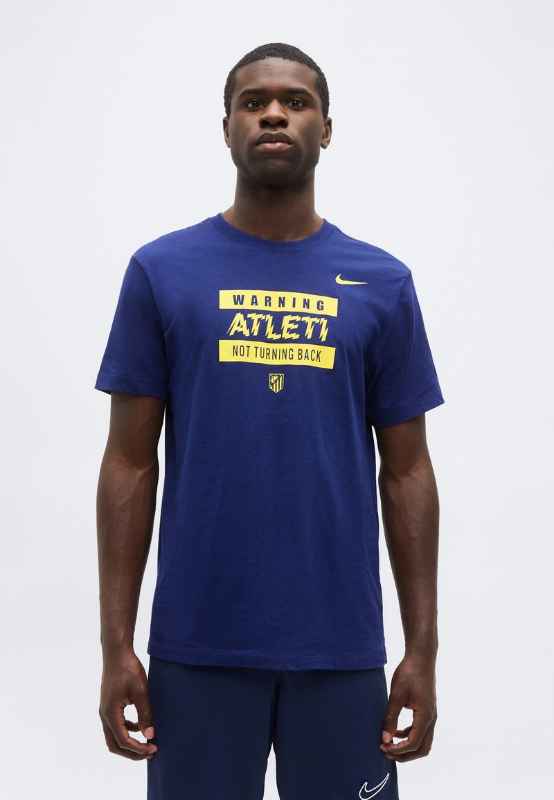 Νεαρός άνδρας που φοράει μπλε μπλούζα Nike με κίτρινο κείμενο "Warning Atleti Not Turning Back" και ταιριαστά μπλε σορτσάκια Nike.