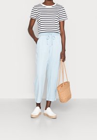 Chemise à manches courtes rayée, pantalon léger bleu clair avec taille élastique, tenant un sac en paille tissé, associé à des baskets blanches.