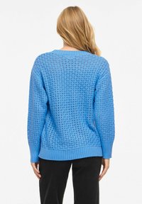 Femme aux longs cheveux blonds portant un pull en laine bleu ample à motif quadrillé et un pantalon noir, vue de dos sur fond blanc.