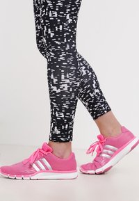 Mustat ja valkoiset kuviolliset legginsit yhdistettynä kirkkaan pinkkeihin urheilujalkineisiin, joissa on hopeiset raidat ja vaaleanpunaiset nauhat, jotka korostavat urheilullista ilmettä.