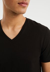Tricou negru cu decolteu în V, realizat dintr-un material moale și elastic, având o textură netedă și un design ajustat, care pune în evidență linia gâtului și gulerul.