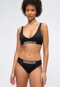 Femme portant un bralette Calvin Klein noir et un slip noir assorti, debout devant un fond clair uni.