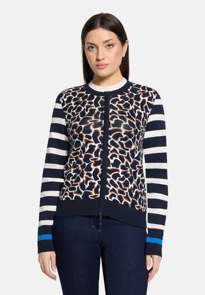 Kobieta ubrana w granatowy sweter na zamek z przodu w panterkę oraz w czarno-białe paski na rękawach z niebieskim mankietem, w połączeniu z ciemnymi jeansami.