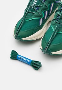 Gr öna sportskor med strukturerat material, flera nyanser och randiga detaljer, i kombination med gröna skosnören och en blå Adidas-märkt etikett.