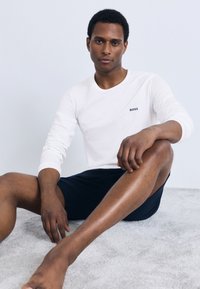 White long-sleeve t-shirt with "BOSS" logo, paired with dark shorts. Model seated on a light gray surface, showcasing clean lines and minimal design.