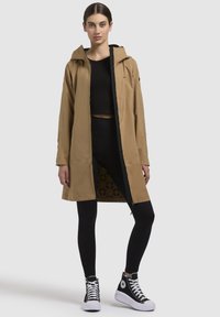 Waterdichte beige lange jas met een capuchon, voorkant met rits en contrasterende zwarte voering; gecombineerd met zwarte leggings en hoge sneakers.