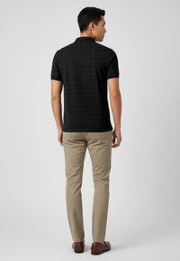 Svart pikétröja med ett subtilt geometriskt mönster, korta ärmar och krage, kombinerad med beige byxor och bruna skor.