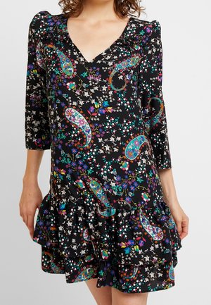 Femme portant une robe noire à col en V avec des motifs floraux colorés et paisley, manches trois-quarts, et un ourlet à volants.