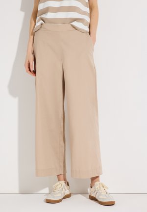 Frau trägt beige weite, cropped Hosen, ein beige-weiß gestreiftes Oberteil und weiße Sneakers mit Kautschuksohlen.