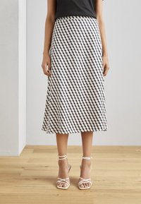 Une jupe mi-longue en A, avec un motif géométrique noir et crème, fabriquée en tissu léger, assortie à des talons blancs à fines lanières.