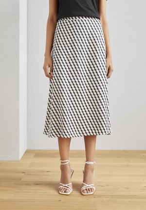 Une jupe mi-longue en A, avec un motif géométrique noir et crème, fabriquée en tissu léger, assortie à des talons blancs à fines lanières.