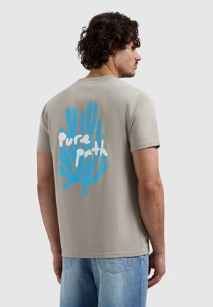 Man met krullend haar die een beige T-shirt draagt met een blauwe koraalgrafiek en witte tekst "Pure Path", gecombineerd met lichtblauwe spijkerbroek.