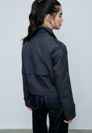 Frau mit dunklem Pferdeschwanz, die eine schwarze, verkürzte Jacke und dunkle Jeans trägt, seitlich vor einem einfarbigen hellgrauen Hintergrund steht.