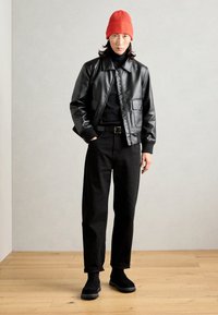 Veste en cuir noir avec un col, col roulé noir, pantalon noir et bottes noires ; accessoirisé avec un bonnet rouge à côtes.