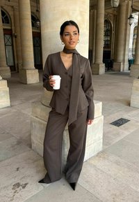 Hnedý upravený oblek so širokými nohavicami a priliehavou bundou, s vyhovujúcim šálom, stojaci pred kamennými stĺpmi. Pohár v ruke.