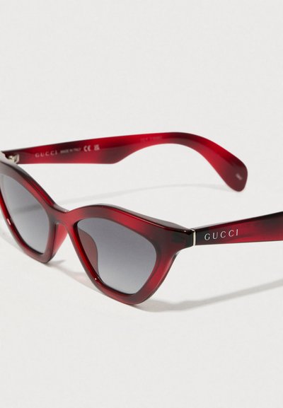 Ochelari de soare din acrilic roșu cu formă de ochi de pisică, lentile gri cu gradient și brandul "GUCCI" în argintiu pe brate. Textură netedă cu finisaj lucios.