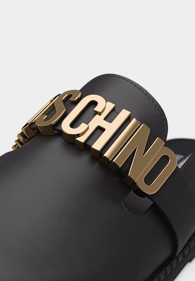 Gros plan d'une sandale noire à glissière avec de grandes lettres dorées "MOSCHINO" sur la lanière.
