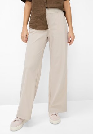 Pantaloni beige a gamba larga indossati con sneakers rosa chiaro e una camicia marrone a maniche corte, in piedi davanti a uno sfondo bianco.