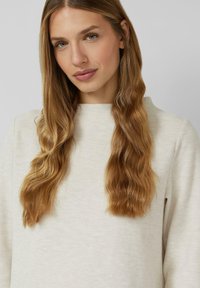 Maglietta a maniche lunghe grigio chiaro a costine con collo alto. Texture liscia, vestibilità comoda, indossata con capelli lunghi, mossi e castani che incorniciano il viso.