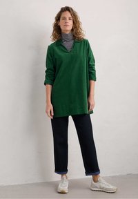 Tunic verde a maniche lunghe con scollo a V, indossata sopra un dolcevita a righe bianco e nere, abbinata a jeans scuri e sneakers bianche.