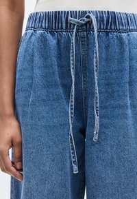 Ανοιχτό μπλε τζιν παντελόνι με ελαστική μέση και κορδόνι για δέσιμο. Το ύφασμα έχει απαλή υφή και διακριτική ξεθώριαση.