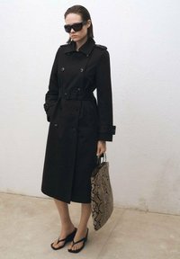 Trench coat negru cu două rânduri de nasturi, epauleți și curea. Purtați cu flip-flops și ținând o geantă din material cu imprimeu de piele de șarpe.