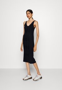 Robe noire tricotée jusqu'aux genoux, dotée d'un décolleté en V, d'une texture côtelée et d'une silhouette ajustée. Associée à des baskets beiges et blanches au style sportif.