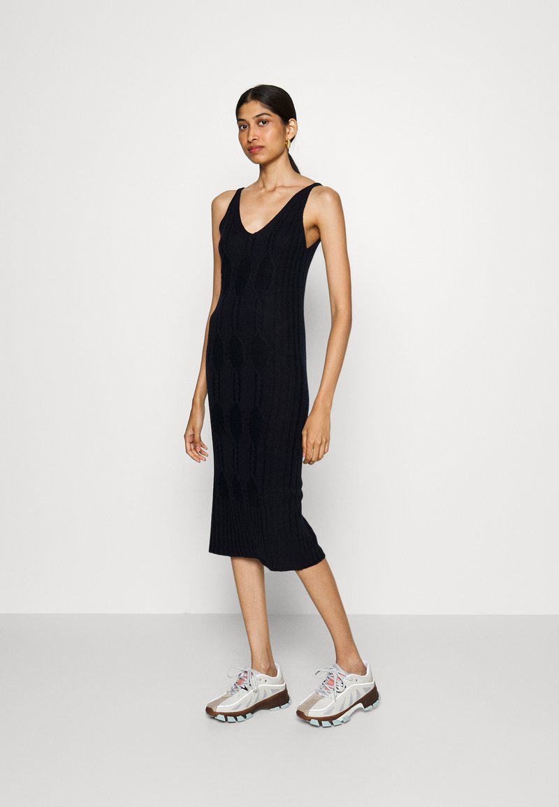 Robe noire tricotée jusqu'aux genoux, dotée d'un décolleté en V, d'une texture côtelée et d'une silhouette ajustée. Associée à des baskets beiges et blanches au style sportif.
