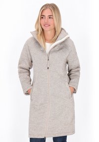 Šviesiaplaukė moteris, dėvinti ilgą, šviesiai rudą zip-up poliarinį švarką su aukštu apykakle, rankas įsidėjusi į kišenes, žiūri šiek tiek į šoną.