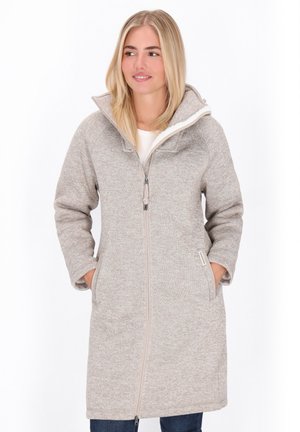 Eine blonde Frau trägt eine lange, beigefarbene Fleecejacke mit Reißverschluss und hohem Kragen. Sie hat die Hände in den Taschen und blickt leicht zur Seite.