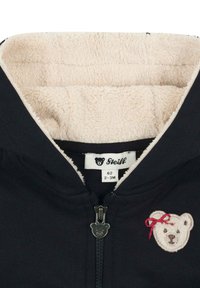 Zwarte ritsjack met een beige fleece voering. Heeft een berenontwerp met een rode strik en een berenvormige ritssluiting. Maatlabel zichtbaar.