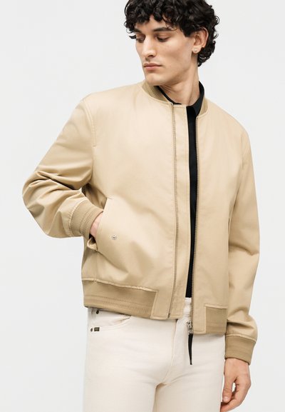 Göndör hajú férfi, bézs bomber dzsekit visel fekete póló felett és fehér nadrágot, lefelé néz, egyik kezét a zsebében tartva.