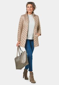 Lichtbeige gewatteerde jas met een hoge kraag, gecombineerd met een witte top, blauwe jeans, grijze suède enkellaarsjes en een metallic grijze schoudertas.