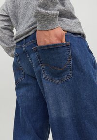 Donkerblauwe denim jeans met een rechte pasvorm, voorzien van achterzakken en contrasterende oranje stiksels. Het materiaal lijkt zacht en versleten.