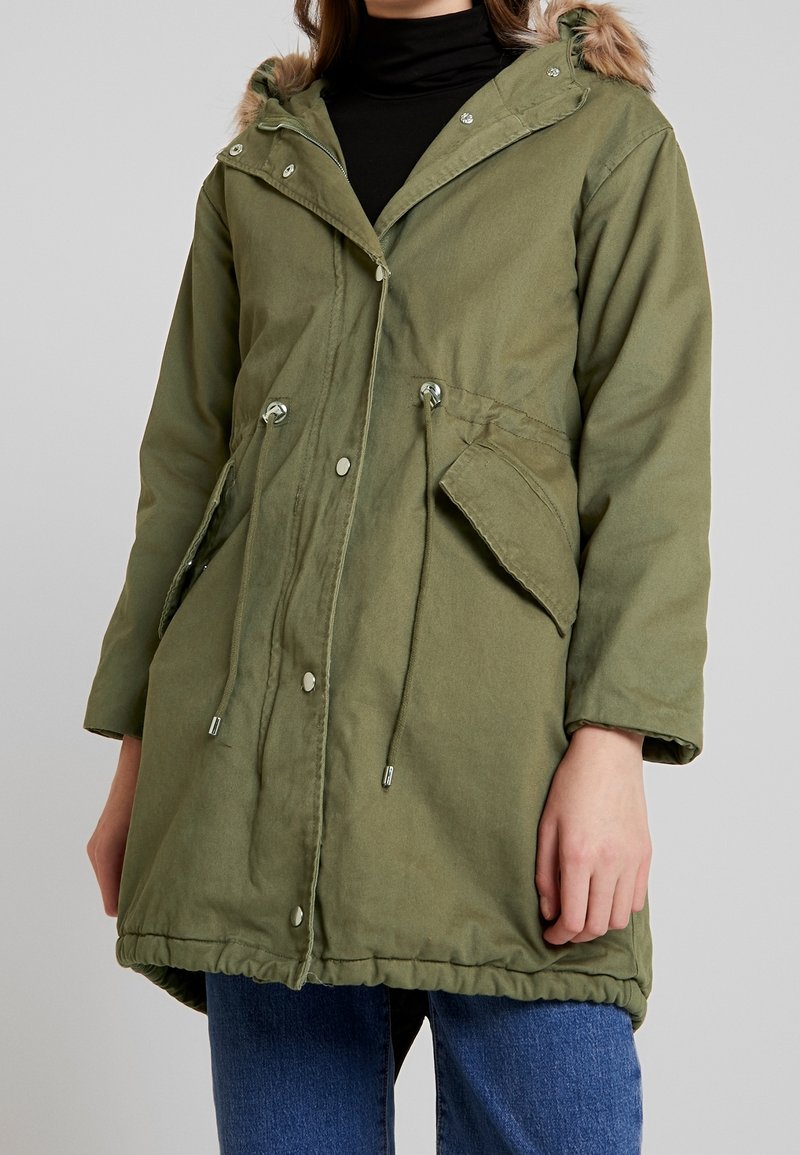 Parka verte avec capuche bordée de fourrure, accessoires argentés, taille à cordon de serrage et poches latérales. Confectionnée dans un tissu durable à la texture douce.