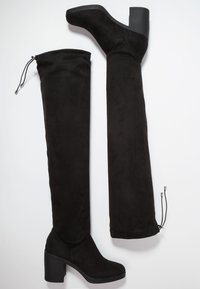 Une paire de bottes montantes noires en daim avec des talons carrés et des cordons ajustables en haut et à l'arrière de l'empeigne.