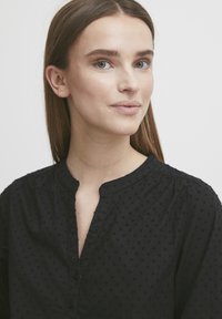Mustmustükk must blouse, millel on tekstuuriline täpikujuline muster, ümar kaelus ja nööp sulgemine. Kude näeb välja pehme ning on pisut vaba lõikega.
