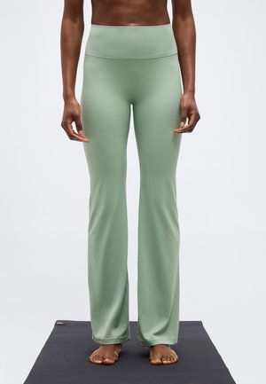 Personne debout pieds nus sur un tapis noir, portant un pantalon taille haute, vert sauge, à jambes larges, visible de mi-torse aux pieds.