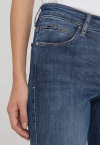 Jeans de mezclilla azul oscuro con un corte clásico, que cuentan con cinco bolsillos, costuras en contraste y un acento de logo metálico en la cinturilla.