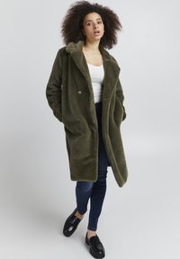 Oliven grøn, fuzzy, dobbeltradet frakke med ærmelapper og to knapper. Sammen med mørke jeans og sorte loafers.