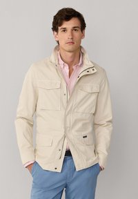 Man draagt een beige jas over een roze overhemd met knopen, handen in de zakken, staand tegen een effen grijze achtergrond.