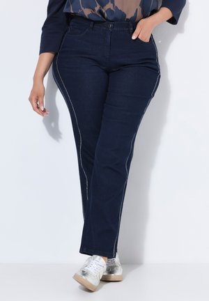 Woman wearing dark blue jeans with white side stitching, metallic silver sneakers, and a patterned navy top, standing against a white wall.