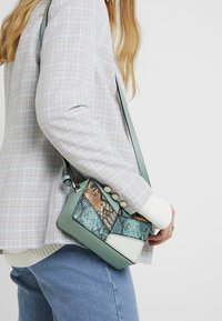 Donna che indossa un blazer scozzese e jeans blu, mentre tiene una piccola borsa a tracolla con un design a pezzi in verde e pelle di serpente.