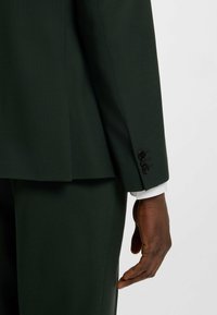 Dunkelgrünes Sakko aus strukturiertem Stoff, mit Schlitz hinten und three-button cuff Detail, unter dem ein weißes Hemd hervorschaut.