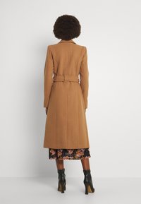 Cappotto trench in misto lana marrone con silhouette aderente, vita cintrata e dettagli sulle spalle. Il cappotto arriva all'altezza del polpaccio.
