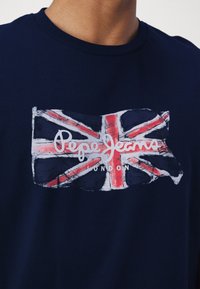 Tamsiai mėlyna medvilninė marškinėliai su nusidėvėjusiu Jungtinės Karalystės vėliavos grafiką; apačioje dizaino yra „Pepe Jeans London“ užrašas baltomis raidėmis.