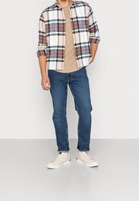 Kostkovaná flanelová košile v krémové, modré a červené barvě přes béžové tričko, doplněná modrými džíny a krémovými teniskami, stojící na neutrálním pozadí.