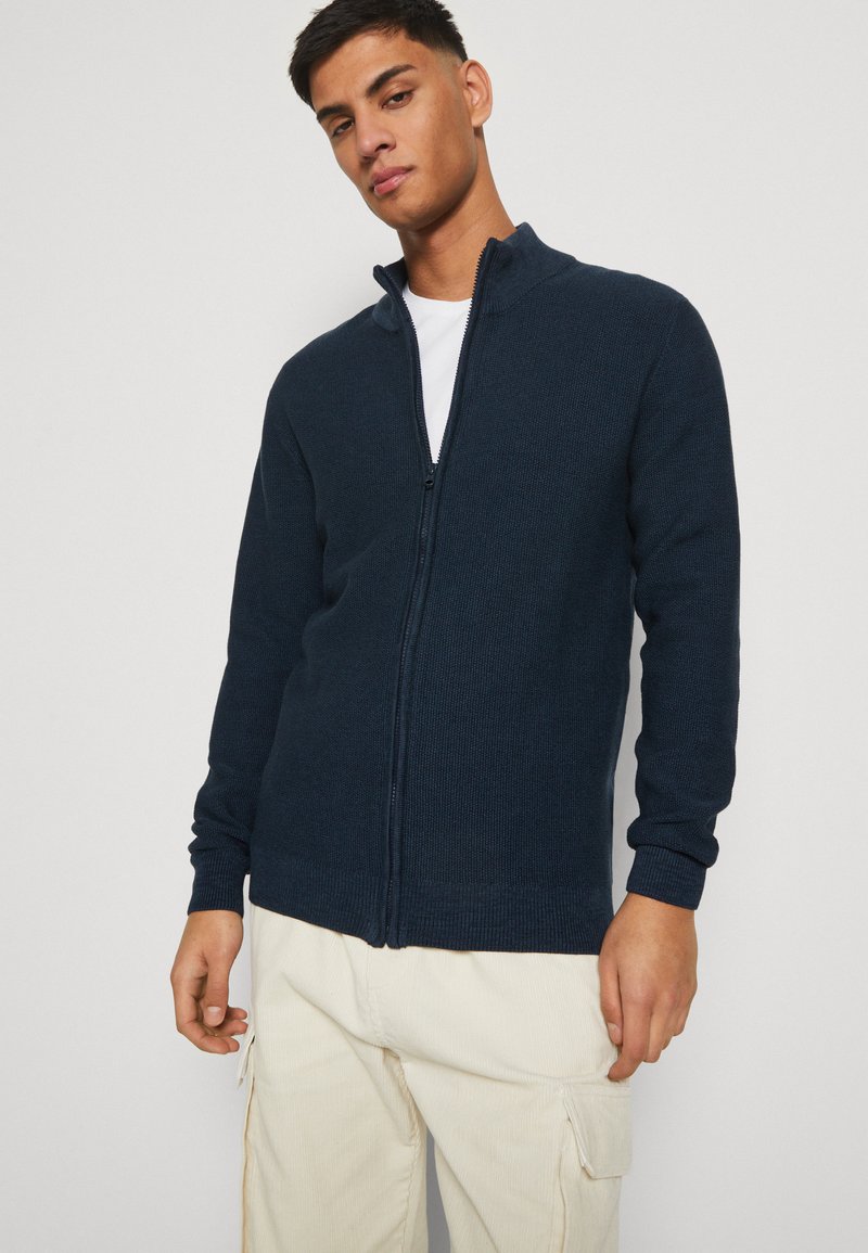 Granatowy sweter zapinany na zamek z teksturowanym splotem, wysokim kołnierzem i ściągaczami. Posiada luźny krój i jest zestawiony z jasnymi spodniami.